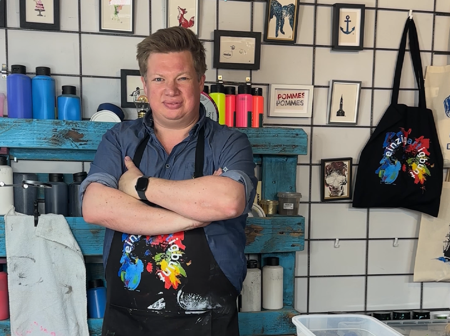 Künstler mit farbenfrohem Schürzen-Design vor Werkstattregal mit nachhaltigen Textilien – einzigartigbunt