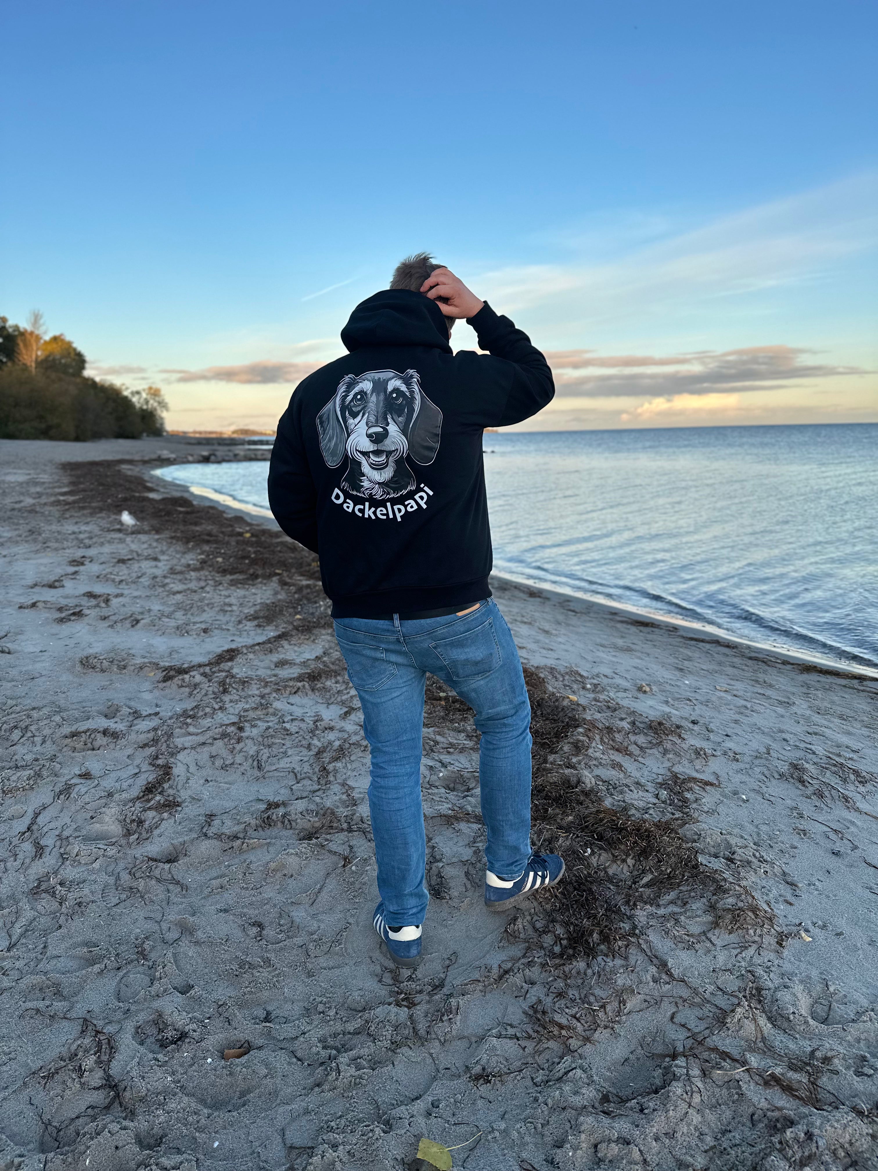 Ein Mensch von hinten am Strand trägt ein Modernen Oversized Hoodie Schwarz in der Rückansicht mit einem großen Print Motiv eines Dackelkopfes und dem geschwungenen Schriftzug Dackelpapi.